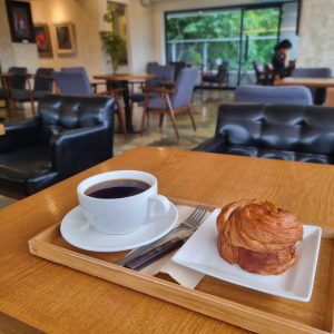 A cup of black coffee and a croissant pastry on a wooden tray, placed on a table inside a cozy café with modern chairs and large windows overlooking greenery