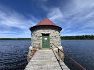 Round stone water tower with a red roof at the end of a wooden walkway over a reservoir.