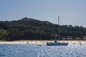 View of the coastline from offshore, with the ocean in the foreground, a sailboat with a motorboat moored to its stern, people on the beach, a building with a bar-restaurant sign, and a lush green hill behind.