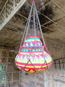 A brightly colored decorative hanging pot, featuring intricate patterns in red, yellow, and blue, suspended from a rope inside a bamboo structure.