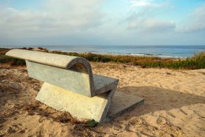 A concrete bench is positioned on a sandy beach, facing the ocean. The bench has a distinctive modern design, with a curved top and a geometric base. In the background, there is a view of the calm sea and a cloudy sky