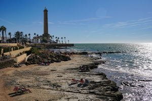 Chipiona, Spain. A coastal scene featuring a lighthouse in the background against a bright blue sky. In the foreground, people are relaxing on the rocky beach and enjoying the sun, while others wade in the water.