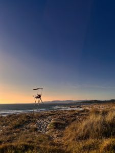 Empty lifeguard chair on a sandy, grassy Atlantic beach at sunset, with the ocean and mountains in the background.