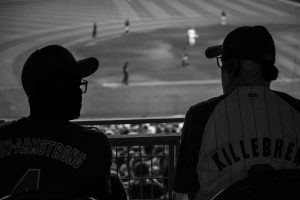 A black and white photo of two men, largely in silhouette, talking while sitting at the top of a section at a baseball game. One is wearing a Chicago Cubs Pete Crow-Armstrong jersey, the other is in a Minnesota Twins Harmon Killebrew jersey