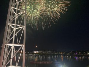 Fireworks along the Ohio River, a reflection of lights in the water. A white tower, a floating boat, and buildings are visible.