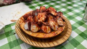 Plate of “pulpo á feira”: sliced octopus drizzled with olive oil and sprinkled with paprika on a round wooden dish, set on a green-and-white checkered tablecloth with a toothpick in the center.