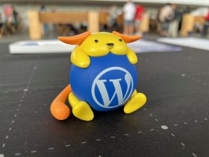 A small yellow Wapuu mascot hugging a blue ball with the white WordPress “W” logo, sitting on a black tabletop with a blurred event crowd in the background.
