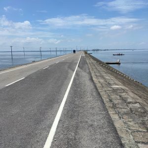 A long, straight road stretching across water on both sides under a bright blue sky, with electric poles lined along the road and small boats visible in the distance. The scene is from Mithamoin, Kishoreganj, Bangladesh