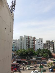 A busy urban street scene. The front row has commercial and multi-story residential buildings of medium to tall heights. The road seems busy with many people engaged in different activities with different vehicles like cycles, motorbikes, rickshaws, and pedestrians.