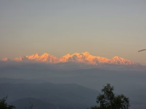 A breathtaking view of snow-capped mountains at sunrise, with the peaks illuminated in soft pink and orange hues. Below, a misty valley stretches out, and patches of greenery can be seen in the foreground.