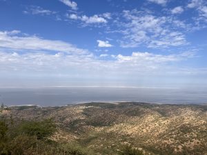 A wide coastal view from a hillside, with dry rolling land, a calm sea, and a blue sky with scattered clouds.