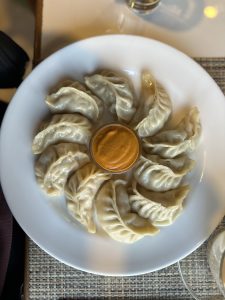 A circular white plate filled with chicken momo arranged in a spiral pattern, surrounding a small bowl of orange dipping sauce in the center.