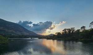 A wide landscape shot of a calm lake reflecting a golden sunset, framed by a large mountain and lush green trees under a soft blue sky.