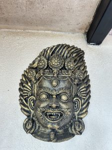 Ornate bronze mask on a wall depicts a fierce, mythological face with wide eyes and bared teeth, adorned with intricate patterns and ornamental details.