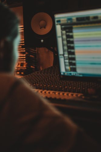 Photographie d'un jeune homme devant un écran affichant des pistes sonores, manipulant les curseurs d'une table de mixage.