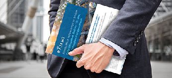 A person in a suit holds documents and a blue RAJANI folder while standing outside in an urban setting with modern buildings in the background.