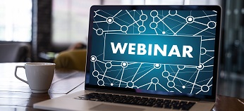 A laptop on a desk displays the word "WEBINAR" on its screen, with a network graphic background. A white coffee cup sits beside the laptop.