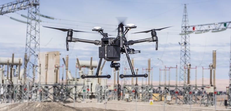 A black drone with multiple rotors hovers in the air at an industrial site, surrounded by electrical equipment, power lines, and transmission towers in the background.