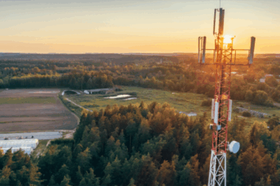 A tall red and white telecommunications tower stands amid a forest and fields at sunset, with the sun glowing in the background and a rural landscape stretching into the distance.