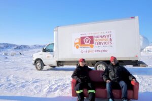 Two men sit on a couch in front of a Nunamoving truck in a snowy landscape