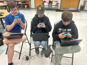 Three students sit in chairs with laptops and their coding devices.