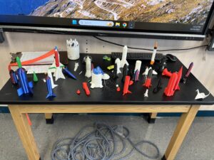 A wide range of 3D-printed rockets sit on top of a classroom table.