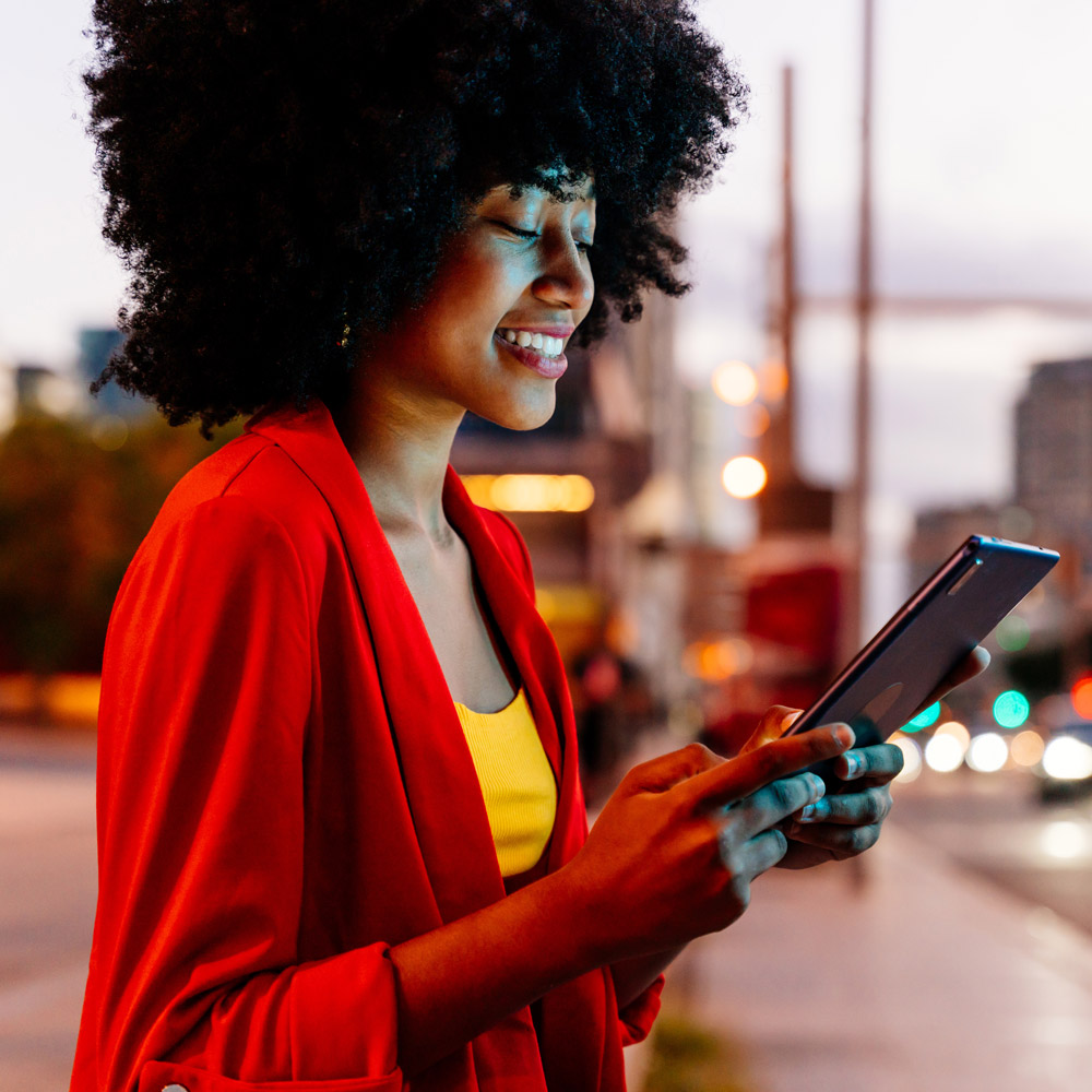 Smiling woman in a red jacket using a tablet on a city street at dusk.
