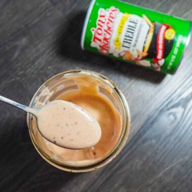 A jar of light orange Raising Cane's dipping sauce with a metal spoon and a jar of Tony Chachere's Creole Seasoning on a gray wooden background.