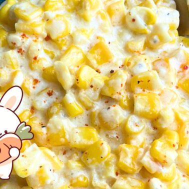 A close-up of a creamy corn dish featuring yellow corn kernels, with a cartoon bunny holding a carrot in the corner.