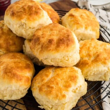 A group of golden-brown buttermilk biscuits is arranged on a round cooling rack, placed on a wooden cutting board. A patterned napkin is partially visible on the right side.