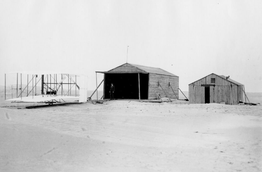 Wright Flyer at the Wright's camp near Kittyhawk, North Carolina. Wilbur Wright is inside the open building near the center of the photograph. (Wright Brothers Aeroplane Company) The Wright's airfield at Kittyhawk, North Carolina. Wilbure Wright is standing in the hangar. (Wright Brothers Aeroplane Company)