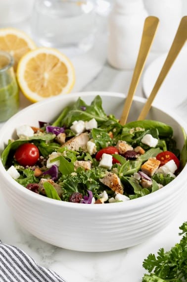 A green salad with tomato, feta, and quinoa in a large white bowl with two gold tone spoon handles sticking out.