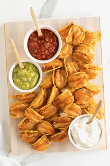 Taco bites made out of tortilla chips, refried beans and cheese served with a bowl of guacamole, sour cream and a bowl of salsa.