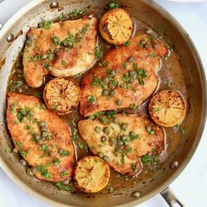 Chicken breast, lemon halves, capers and sauce in a skillet with a white bowl filled with brown rice and green peas and a white plate with toasted baguette slices on it.
