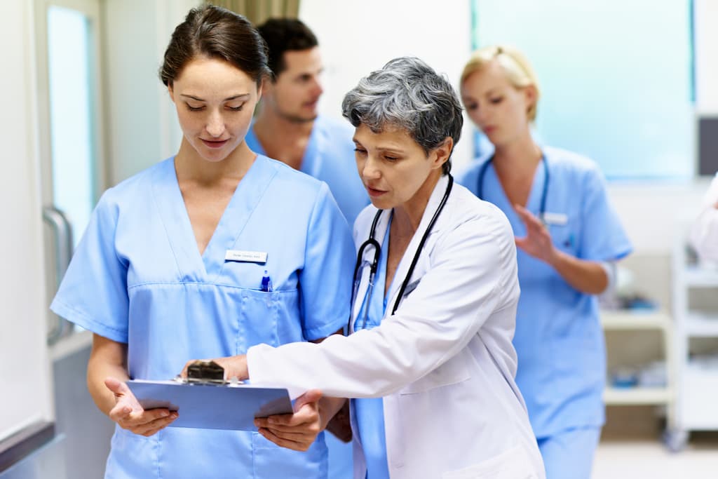 A doctor in a white coat holds a clipboard while two nurses in blue scrubs stand nearby in a hospital room.
