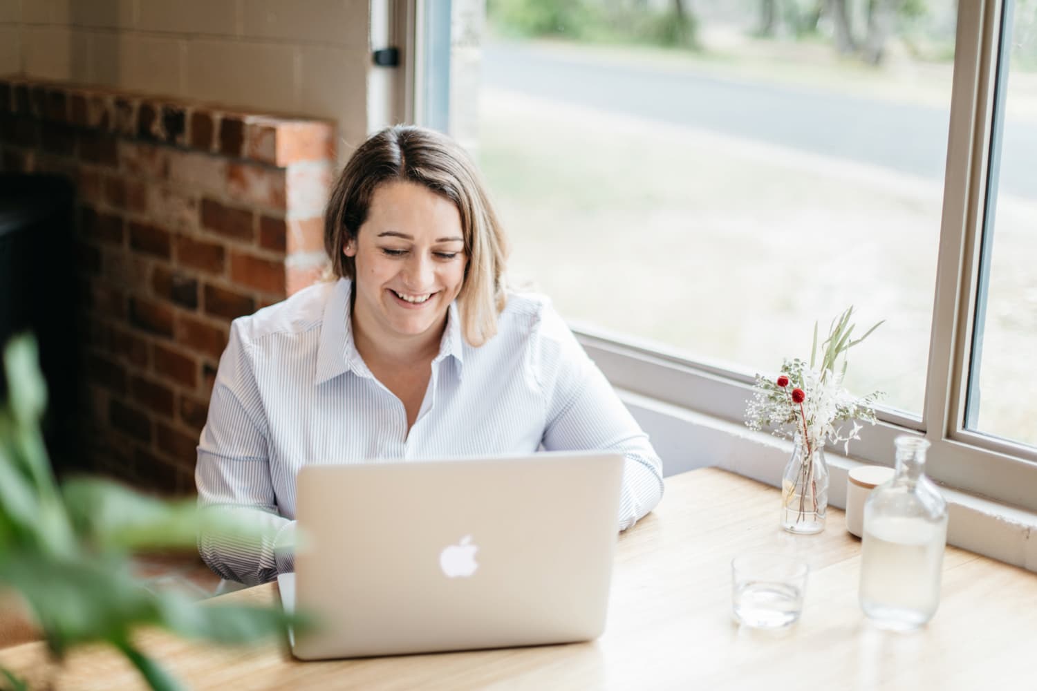The Island Creative - Erica on Computer A person with shoulder-length hair sits at a wooden table by a large window, smiling while using a silver laptop. A glass of water, a bottle, and a small vase with flowers are on the table.