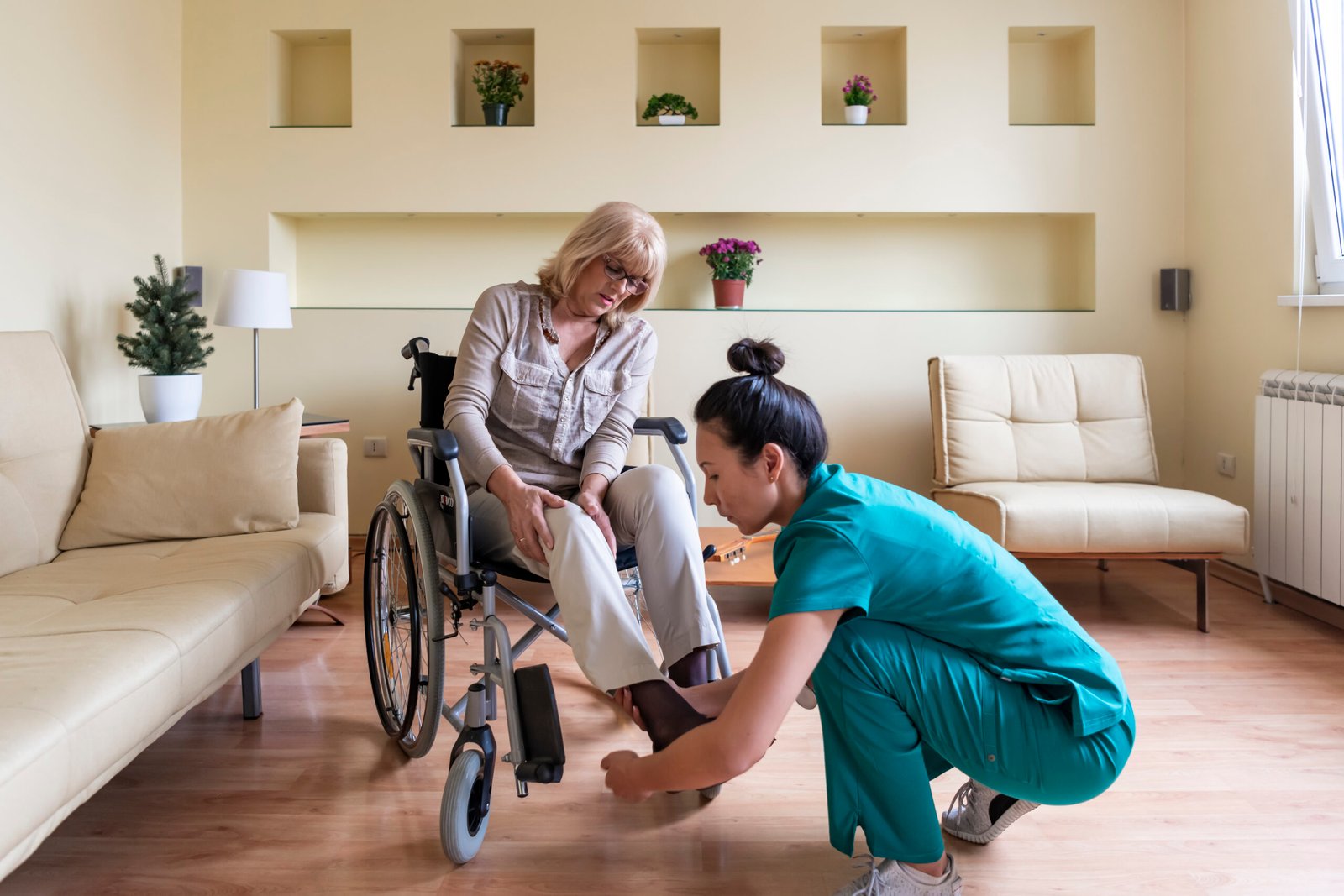 Sick Old Woman in a Wheelchair is Receiving a Help From a Young Nurse in Home Visit. The Learning Company - Health Care Aide