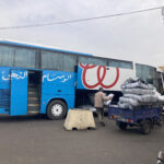 Un bus bleu est stationné, avec la porte passagers ainsi que la soute ouvertes. Un homme de dos, qui porte un casque de moto sur la tête, est en train de mettre de grands sacs blancs à l'intérieur de la soute, qui en contient déjà quelques uns. A ses côtés se trouve un triporteur bleu rempli des mêmes sacs, qu'il est en train de décharger. Ce bus est collé contre un autre bus dont on ne distingue que l'arrière