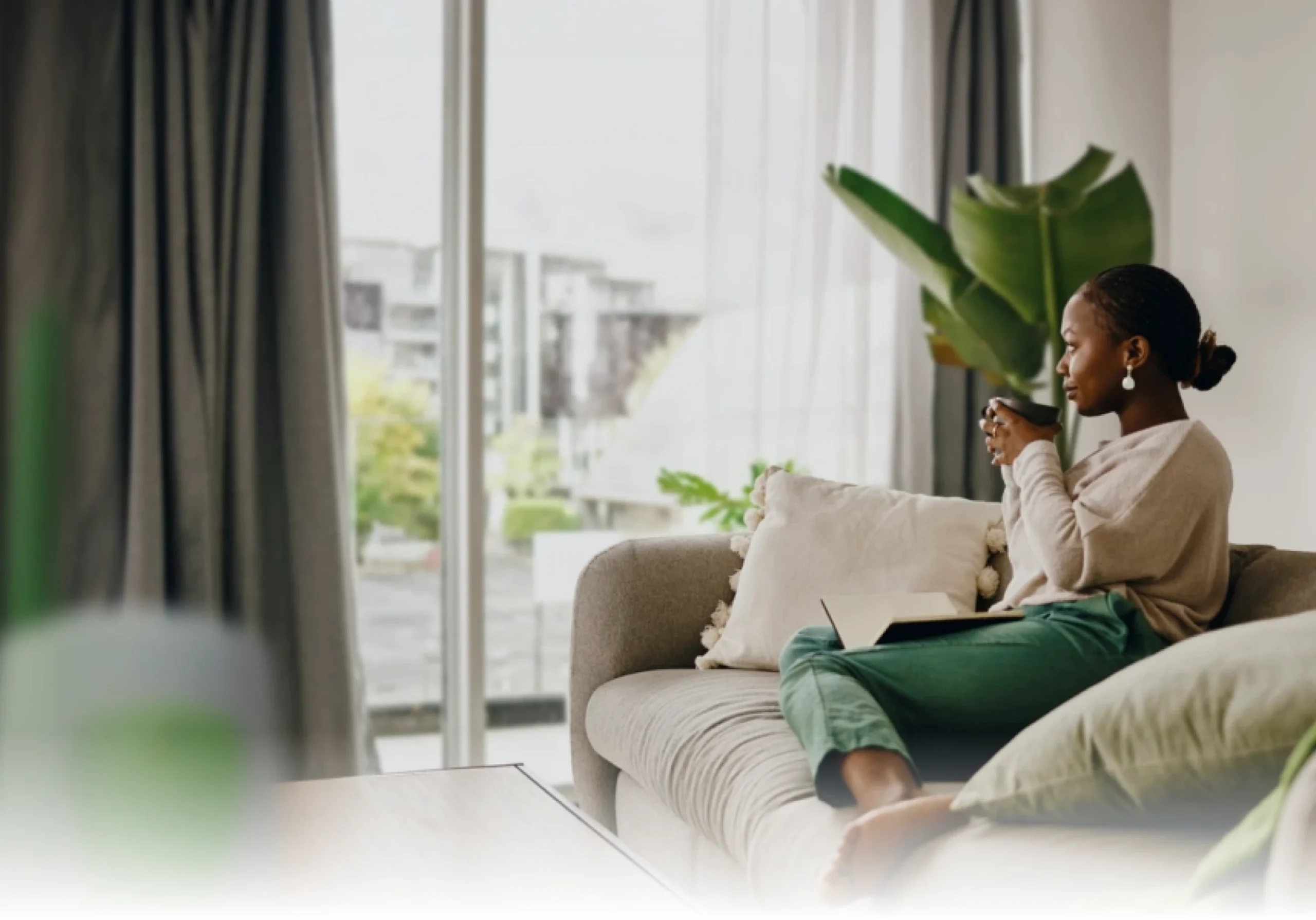 A woman sits on a sofa by a large window, holding a mug and looking outside. She has a laptop on her lap, surrounded by cushions and indoor plants, with natural light filling the modern living room.