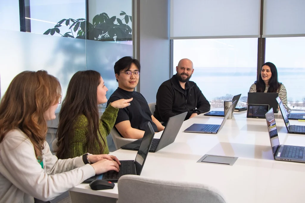 Five people sit around a conference table with laptops, engaged in discussion. Large windows and a plant are in the background, giving the room a bright, modern atmosphere.