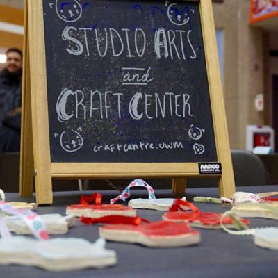 Table display featuring a chalkboard sign that reads 'Studio Arts and Craft Center' with the URL 'craftcentre.uwm' written below. The sign is decorated with small doodles. In front of the sign, several handmade craft items with colorful ribbons are arranged on the table.