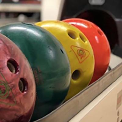 Close-up of four colorful bowling balls arranged in a rack. The balls are red, green, yellow, and orange, each with three finger holes and visible logos. The background shows part of a bowling lane and ball return system.