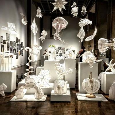 Art gallery display featuring illuminated white paper sculptures in various abstract and organic shapes, including stars, spirals, jellyfish-like forms, and geometric designs. The sculptures are arranged on white pedestals and suspended from the ceiling, creating a glowing, ethereal atmosphere. Framed artwork is visible on the wall in the background, and visitors are observing the exhibit.
