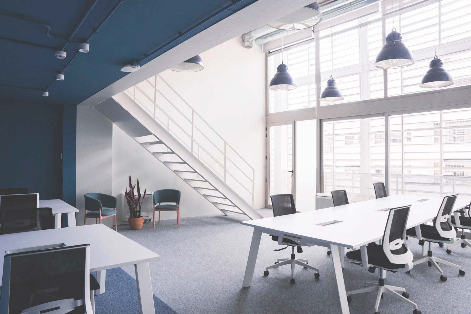 Mesa de oficina blanca con sillas, escaleras y lámparas colgantes, en oficina moderna.