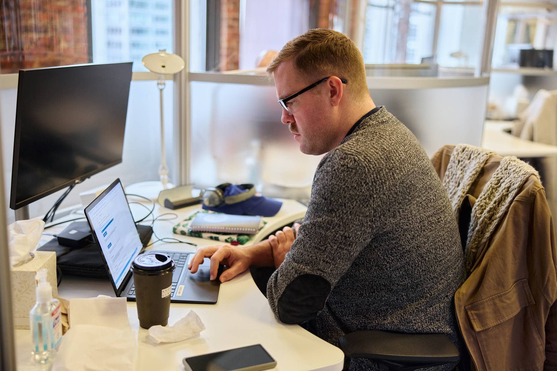 A man working at a desk in an office, using a laptop with a black monitor nearby, surrounded by a coffee cup, hand sanitizer, and notebooks.