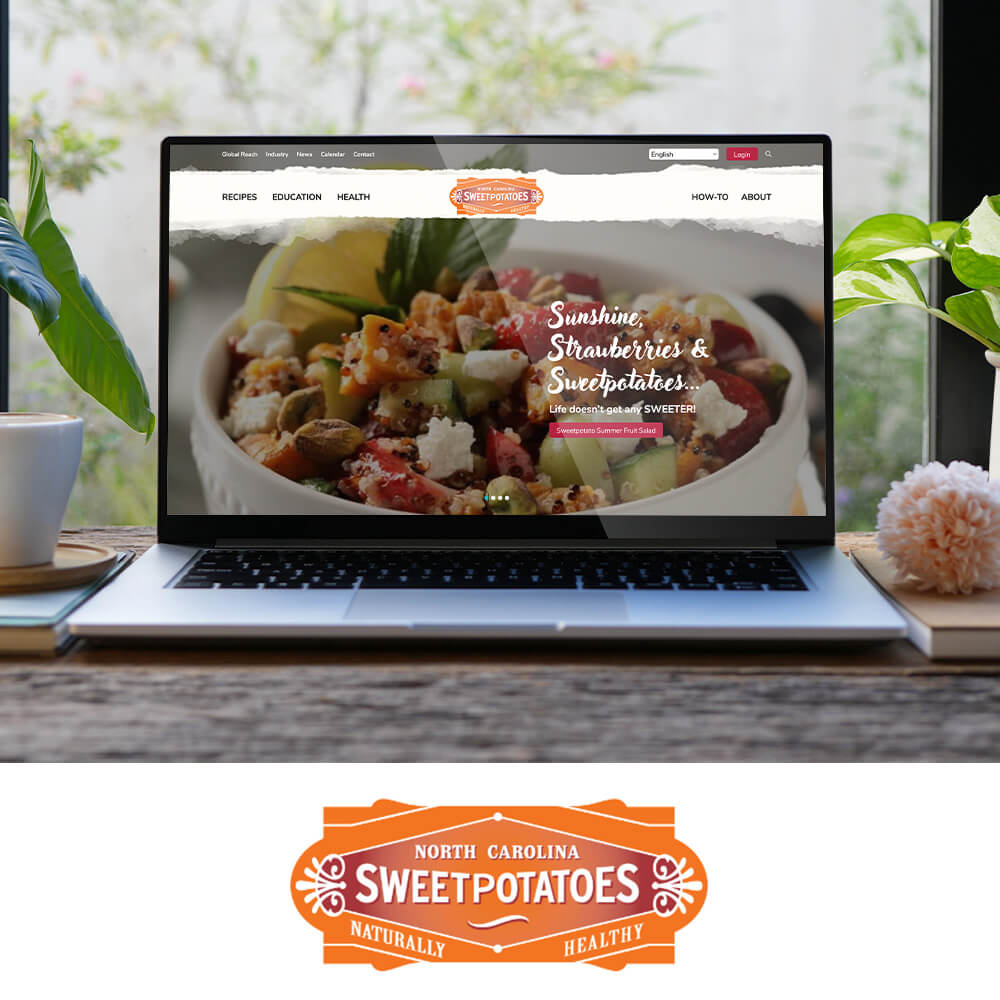 A laptop on a wooden desk displays the North Carolina Sweetpotatoes website. The homepage shows a large bowl of fruit and sweetpotato salad with text overlay: “Sunshine, Strawberries & Sweetpotatoes… Life doesn’t get any SWEETER!” A pink button below reads “Sweetpotatoes Summer Fruit Salad.” The navigation bar includes links for Recipes, Education, Health, How-To, and About. Plants, a coffee cup, and a notebook sit beside the laptop. Below the main photo is the North Carolina Sweetpotatoes orange logo with the words “Naturally Healthy.”