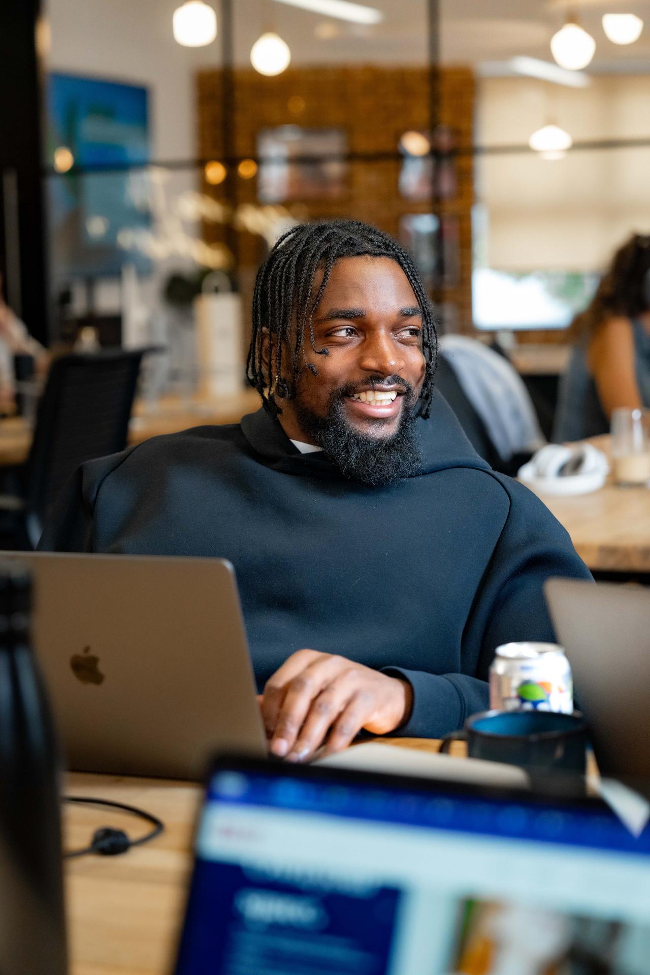 A cheerful man in a black hoodie, with braided hair and a beard, enjoys working on his laptop in a bright office with exposed brick walls and glass partitions.