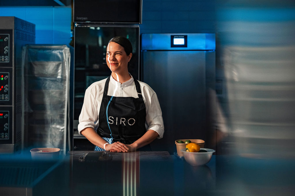 Adult woman in her 30s with dark hair pulled back stands behind a kitchen counter, wearing a white chef’s jacket and black apron, smiling calmly inside a professional kitchen.