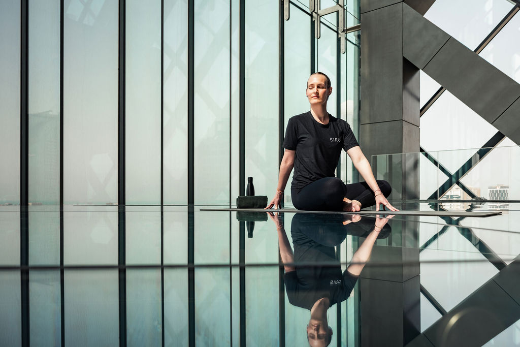 Adult woman in her 30s with dark hair tied back sits cross-legged on a yoga mat, wearing black activewear, practising meditation inside a modern studio with floor-to-ceiling glass walls.