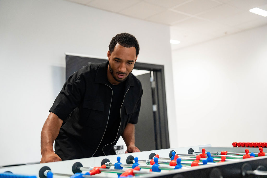 Adult man wearing a black short-sleeve shirt leans over a foosball table, concentrating on the game in a bright office breakout space with white walls and overhead lighting.